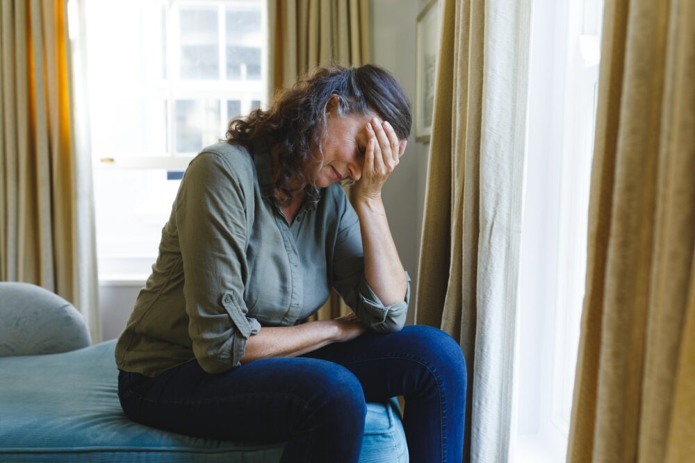Stressed senior caucasian woman sitting on sofa by window holding head